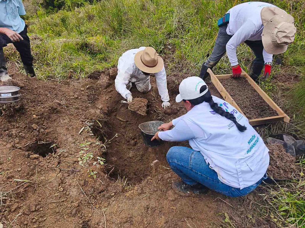 Primer plano de tres personas del equipo humanitario trabajando en el suelo: una persona excavando con palustre, otra recolectando tierra en un balde y otra tamizando el terreno con un cernidor de madera.
