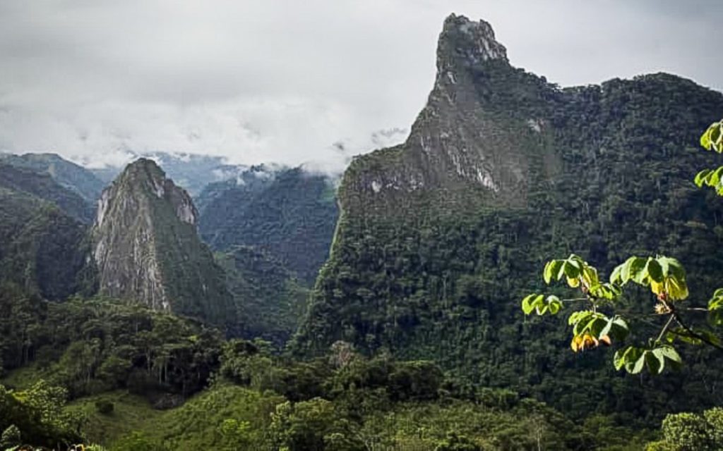 Vista panorámica de un relieve montañoso cubierto por boscosa vegetación tropical. Destaca una alta formación rocosa y empinada en el centro-derecho y otra menor a la izquierda, rodeadas de neblina baja y nubes en el horizonte.