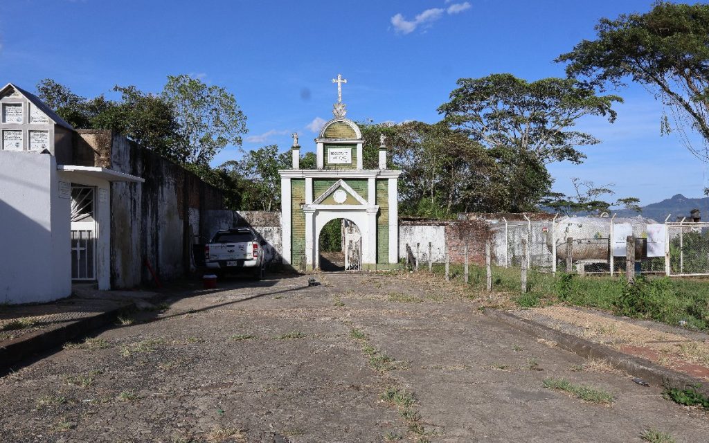 Fachada de la entrada a un cementerio en zona rural. Presenta un portal de color blanco con acabados verdes, columnas y una cruz en la parte superior. A la izquierda se observa una edificación blanca con nichos y una camioneta pick-up blanca aparcada. Al fondo se divisan árboles y montañas bajo un cielo despejado.
