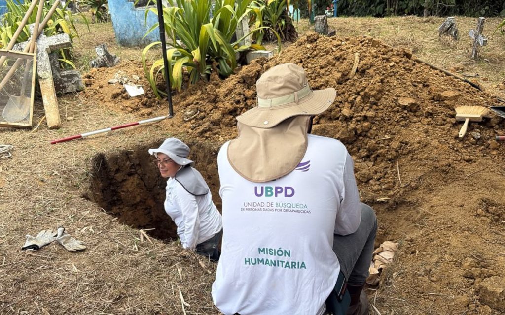 En primer plano, la espalda de un especialista en misiones humanitarias con camiseta blanca que lleva el logo de la UBPD y sombrero de ala ancha, supervisando a una compañera situada dentro de una fosa en el suelo rodeada de montículos de tierra y herramientas de excavación.