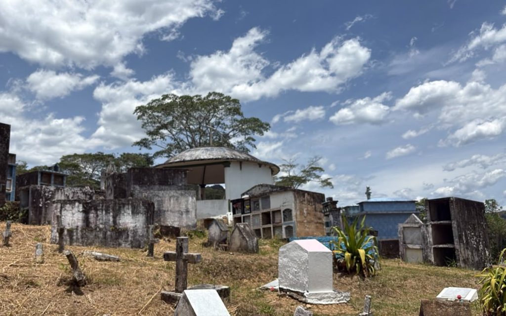 Vista abierta de un cementerio sobre una colina con tumbas dispersas, lápidas blancas y cruces de piedra o cemento. En la parte central sobresale una estructura circular blanca tipo kiosco o templete, bajo un cielo azul con densas nubes blancas.