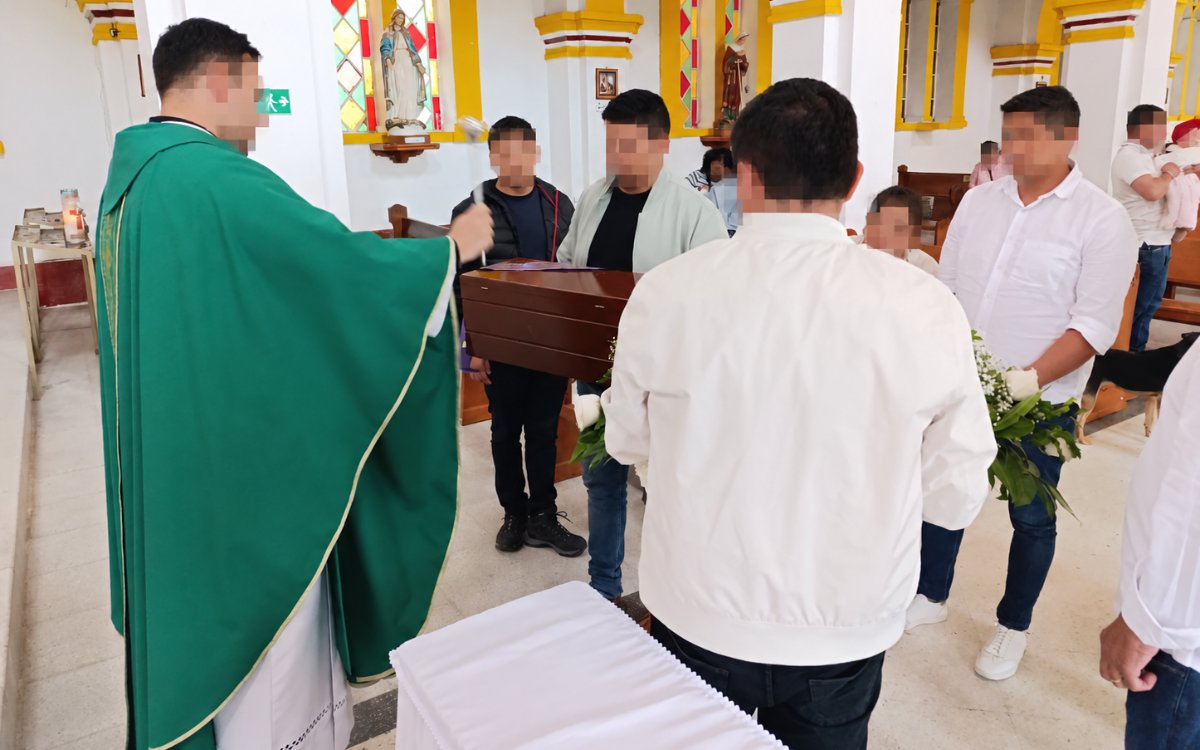 Fotografía tomada en el interior de un templo católico. A la izquierda, un sacerdote de espaldas viste una casulla verde y sostiene un hisopo con el que rocía agua bendita hacia el centro. Frente a él, un grupo de hombres sostiene un cofre fúnebre de madera oscura cubierto parcialmente. La nave del templo tiene paredes blancas con detalles en amarillo y vitrales al fondo.
