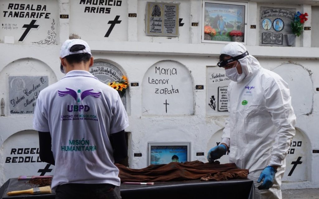 Dos especialistas de la UBPD, uno de ellos portando traje de bioseguridad blanco y mascarilla, observan minuciosamente y organizan restos biológicos sobre una mesa negra en las galerías de un cementerio con nichos sepulcrales blancos de fondo.