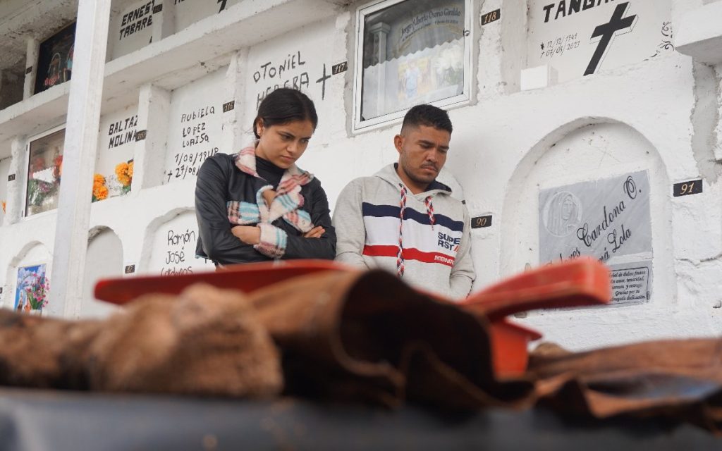 Plano medio de una joven con los brazos cruzados y un hombre a su lado en el cementerio, observando hacia abajo con semblante serio durante la labor de la misión humanitaria.