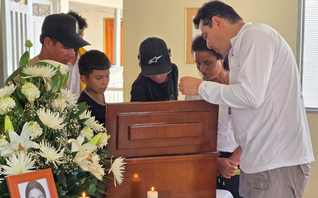 Un grupo de familiares, incluyendo niños y adultos, permanecen de pie con la cabeza inclinada en señal de respeto alrededor del cofre de madera de su ser querido. A la izquierda se observa un arreglo floral con flores blancas.