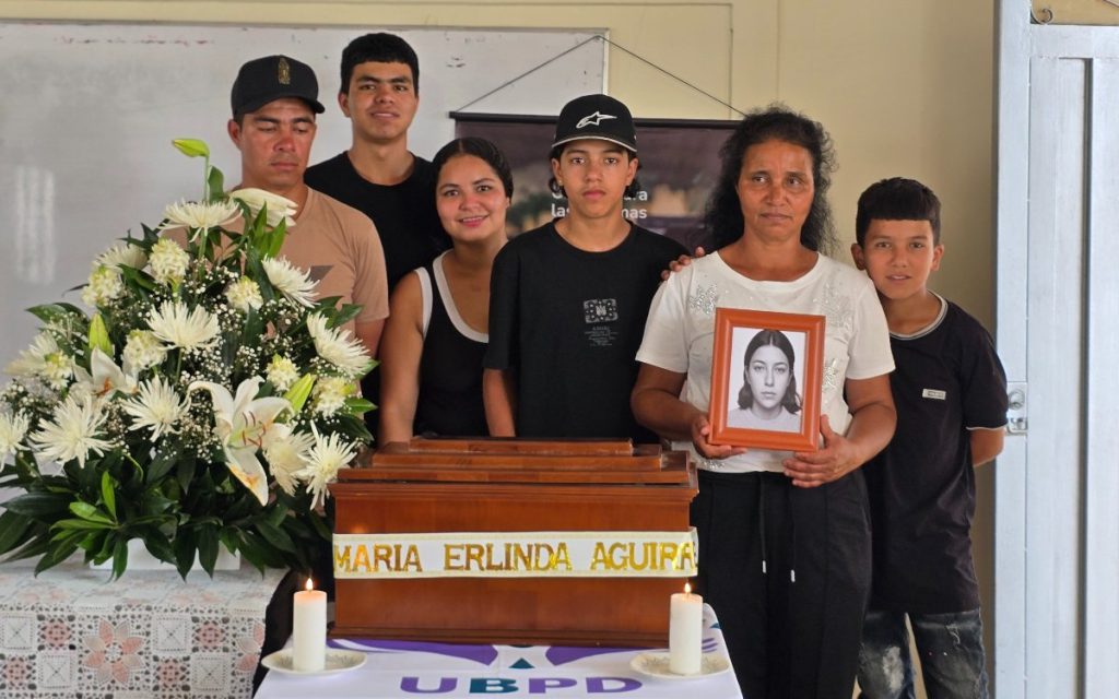 Seis integrantes de una familia de pie detrás de una mesa cubierta con un mantel con las siglas UBPD, sobre la cual descansa el cofre de madera escoltado por dos velas encendidas. Una mujer sostiene en sus manos el retrato enmarcado en madera de la persona fallecida.