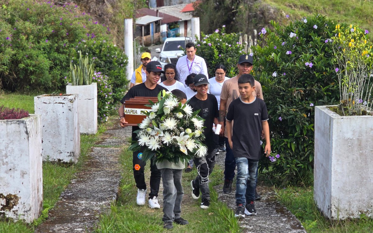 Varios familiares caminan en procesión por un camino de cemento rodeado de vegetación y jardines en un cementerio. En la parte delantera, dos jóvenes transportan el cofre fúnebre y una gran corona de flores blancas hacia el lugar de sepultura.