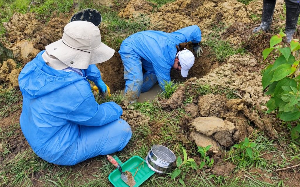 Plano en picado de dos integrantes del equipo técnico forense vistiendo trajes e impermeable azul. Una persona se encuentra agachada examinando la tierra dentro del pozo excavado, mientras la otra permanece sentada a un costado asistiendo con un cernidor metálico y una pala pequeña sobre el terreno pastoso.
