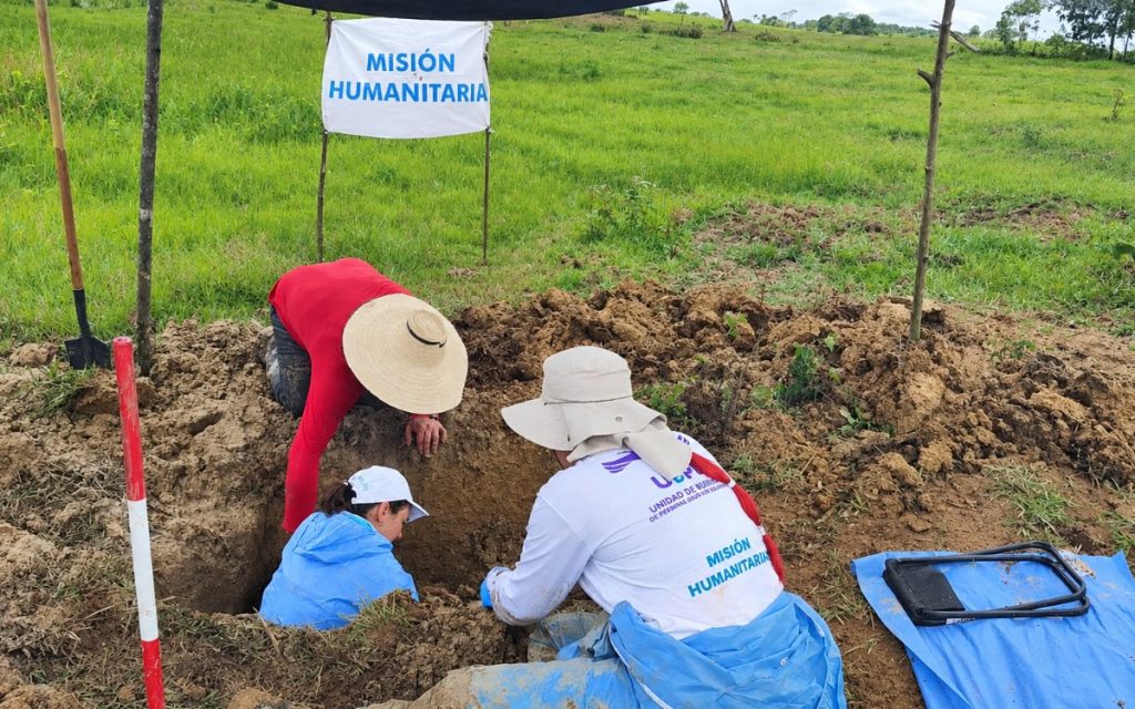 Integrantes del equipo forense de la Unidad de Búsqueda de Personas dadas por Desaparecidas (UBPD) trabajando en un punto de excavación en el campo. Al fondo se observa un cartel blanco sujeto a dos estacas con el texto en letras azules "MISIÓN HUMANITARIA", mientras los profesionales trabajan agachados y sentados junto al orificio cavado en la tierra.