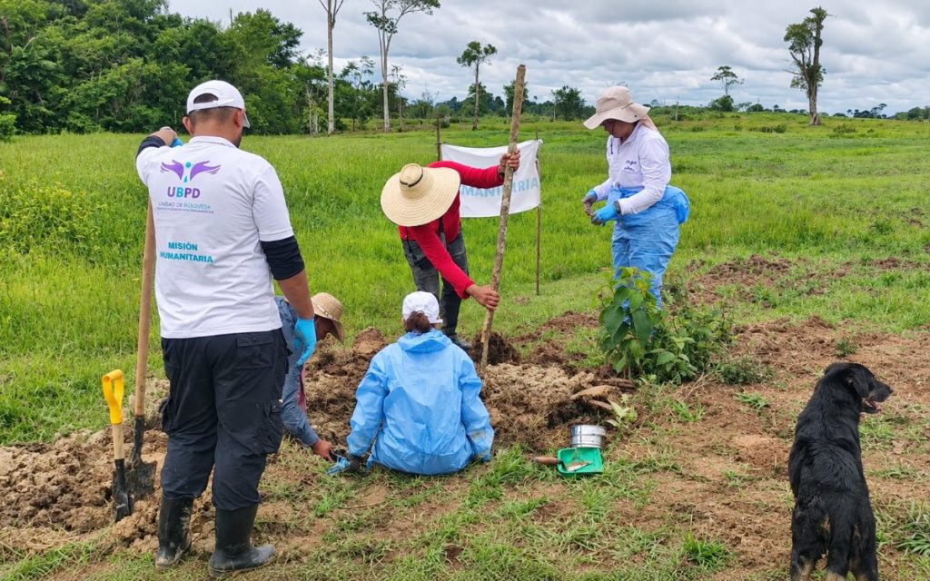 Vista panorámica de un grupo de cinco personas realizando labores de excavación en un terreno verdoso de campo. Un profesional viste la camiseta con el logo de la UBPD y el texto "Misión Humanitaria" de espaldas a la cámara. En primer plano a la derecha, un perro negro observa la escena mientras el equipo manipula palas, cernidores y herramientas de prospección.