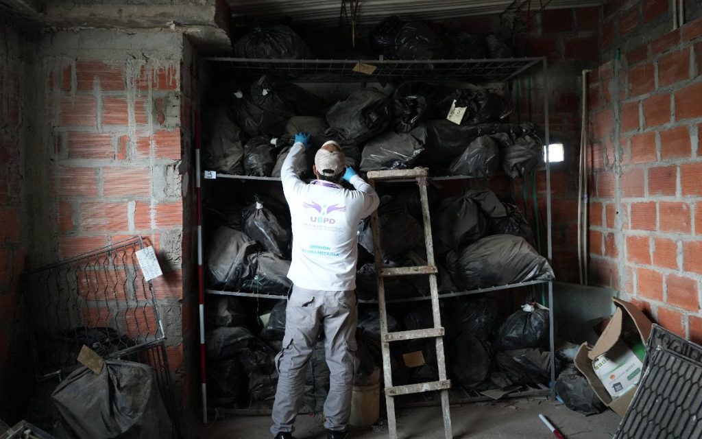 El funcionario de la UBPD, de espalda y usando guantes azules de protección, alcanza con las manos la parte alta de la estantería llena de bolsas negras. A su lado hay una escalera de madera apoyada contra la estructura en un recinto de ladrillo.
