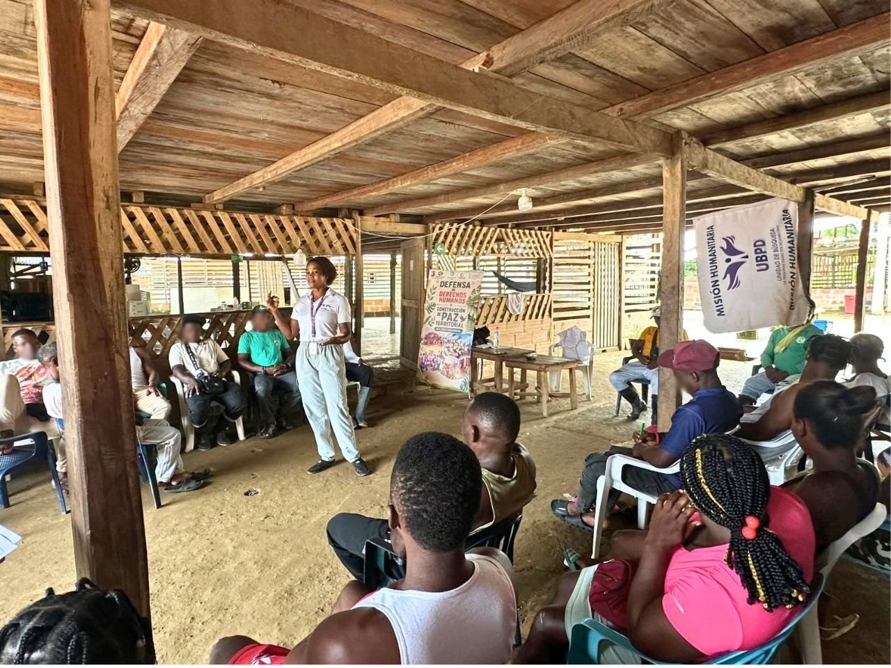 Reunión comunitaria en un espacio abierto con estructura y piso de madera. Al centro, una profesional que viste camiseta blanca con distintivo de la Unidad de Búsqueda de Personas dadas por Desaparecidas (UBPD) habla de pie hacia un grupo de habitantes sentados a su alrededor. Al fondo, un pendón institucional dice 'Defensa de los Derechos Humanos, Construcción de Paz Territorial' y un pendón de 'Misión Humanitaria'.