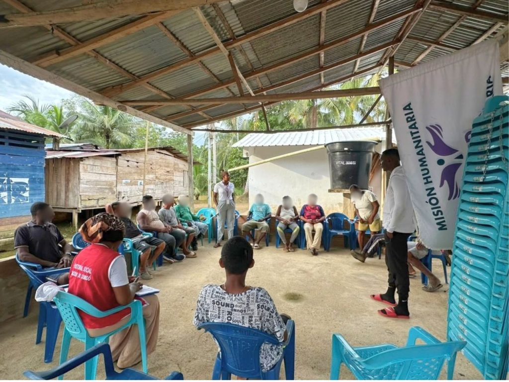 Toma amplia de una reunión participativa al aire libre bajo una cubierta de lámina y vigas de madera. En el centro, una mujer de blanco y, a la derecha, un integrante de la misión humanitaria conversan con un grupo de residentes sentados en sillas plásticas. A la izquierda, una persona lleva un chaleco rojo con la inscripción 'Pastoral Social'.