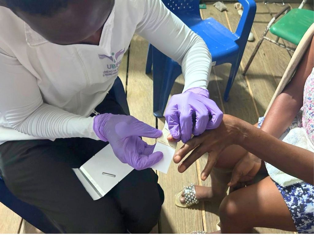 Plano cerrado superior que muestra las manos de una funcionaria de la UBPD utilizando guantes nitrilo morados para presionar suavemente la huella o muestra de sangre del dedo de una persona sobre una tarjeta de recolección de muestras biológicas sobre un sobre de papel blanco.