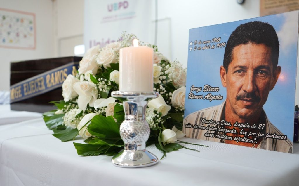 En primer plano, una vela blanca encendida sobre un candelabro metálico adorna un arreglo de flores y rosas blancas. A la derecha, un retablo impreso muestra el rostro de Jorge Eliécer Ramos Algarín, con sus fechas de nacimiento y fallecimiento (2 de enero 1965 – 5 de abril de 1999) y un mensaje conmemorativo familiar. Al fondo se aprecia un cofre funerario de madera con una cinta azul con el nombre en letras doradas.