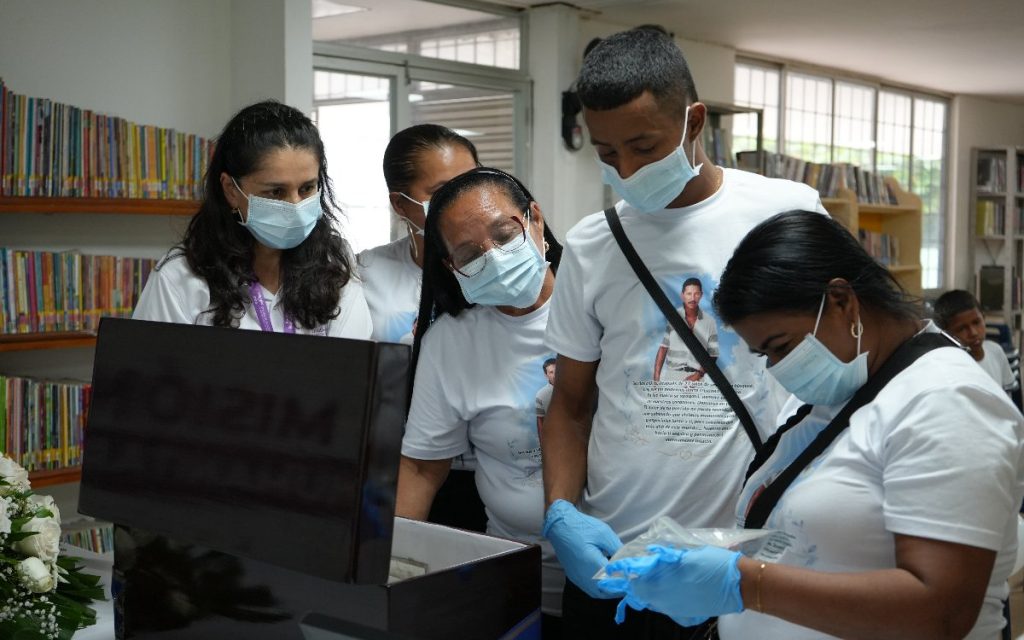 Un grupo de personas, entre ellos hombres y mujeres vistiendo camisetas blancas con la fotografía conmemorativa de su familiar y tapabocas, se inclinan sobre un cofre de madera para revisar bolsas con evidencias dentro de un espacio institucional.