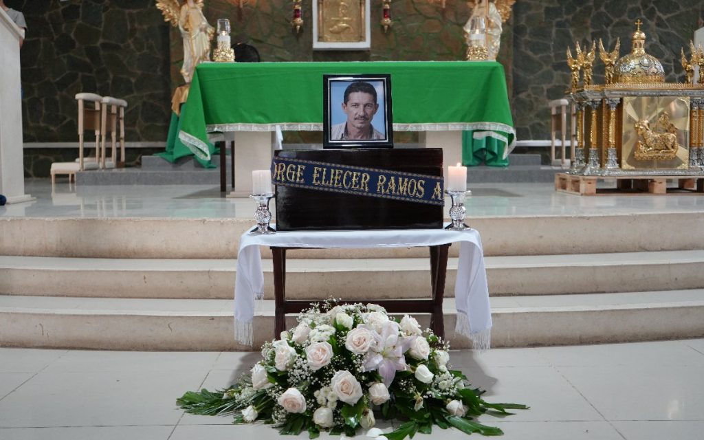 En el centro de la nave principal de un templo o iglesia católica, un cofre de madera está posado sobre una mesa con mantel blanco. En la cinta azul sobre el cofre se lee "Jorge Eliécer Ramos A.". Encima del cofre descansa la fotografía enmarcada del fallecido y a los lados hay velas encendidas. Un arreglo florar blanco decora la base en el piso.