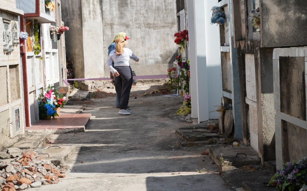 Dos personas en un pasillo de cementerio entre bóvedas blancas, observando un área acordonada con una cinta rosada.