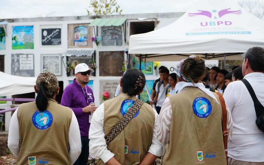 Vista desde atrás de un grupo de personas, entre ellas mujeres vistiendo chalecos beige con logotipos de la UBPD y el municipio de Aipe. Al frente, un funcionario con camiseta morada y gorra blanca habla con el grupo. Al fondo se observa una carpa blanca institucional de la UBPD y la estructura de bóvedas del cementerio.