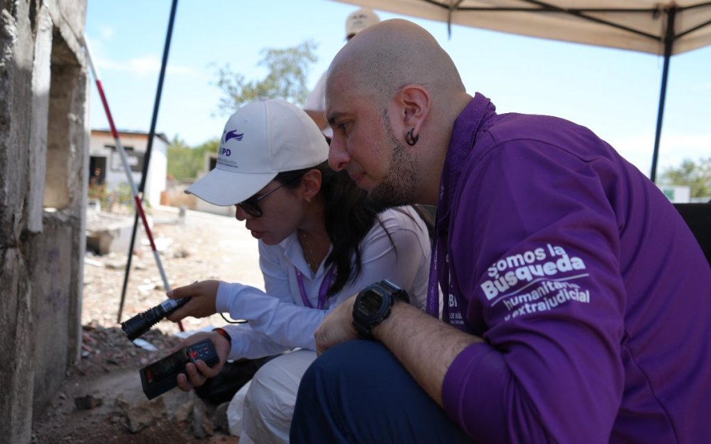Plano medio de dos personas agachadas en labor técnica: una mujer con gorra y camisa blanca apuntando con una linterna hacia una pared de cemento, y a su lado, un hombre con camiseta morada que lleva impreso el lema "Somos la búsqueda humanitaria y extrajudicial", observando detalladamente la medición.