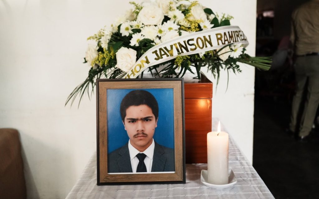 Retrato enmarcado del joven Jhon Jaivinson Ramírez sobre una mesa con una vela encendida, delante de un pequeño cofre fúnebre de madera adornado con flores blancas y una cinta con su nombre.