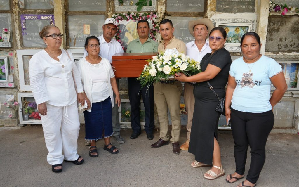 Grupo de seis familiares en un cementerio, de pie frente a un muro de osarios. Dos hombres sostienen en el centro el cofre fúnebre de madera y una mujer sostiene el arreglo de flores blancas.