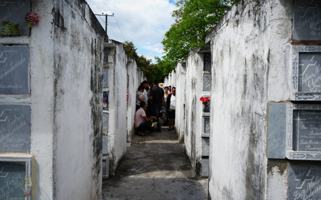 Pasillo estrecho entre dos muros blancos llenos de osarios en un cementerio, con un grupo de personas reunidas al fondo participando en la ceremonia.