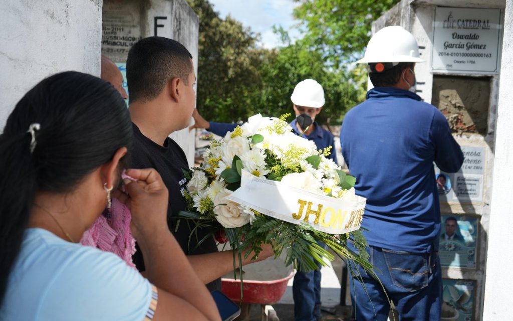 Un hombre sostiene el arreglo floral de Jhon Jaivinson mientras observa a dos trabajadores del cementerio sellar el osario. En primer plano, una mujer se lleva la mano al rostro, conmovida.