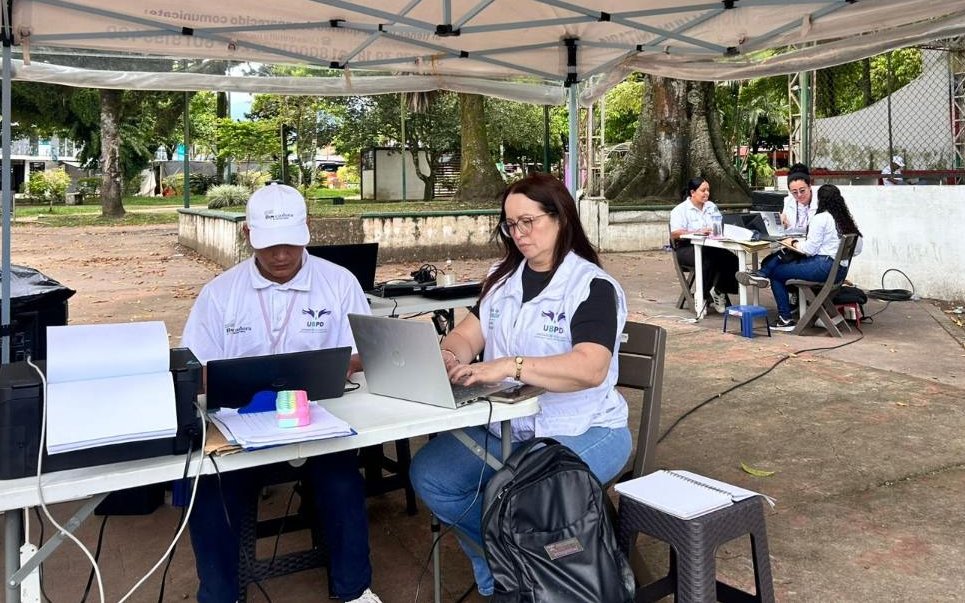 Dos profesionales de la UBPD sentados frente a una mesa plegable trabajando en computadores portátiles bajo una carpa blanca de atención. Al fondo, otros compañeros conversan y registran datos de usuarios en un entorno de parque o plaza.