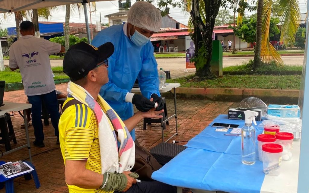 Primer plano de una toma de muestra biológica en campo. Un profesional de la UBPD equipado con bata médica azul, gorro, tapabocas y guantes realiza una punción capilar en el dedo de un ciudadano con camiseta amarilla para la recolección de ADN. Sobre la mesa hay frascos de muestra, antiséptico y material médico.