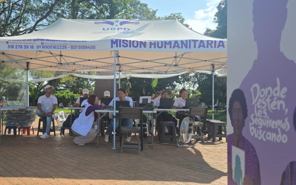 Vista general del stand institucional al aire libre bajo una carpa que dice "UBPD - Misión Humanitaria". En primer plano a la derecha resalta un pendón morado con el texto "Donde estén, les seguiremos buscando" y teléfonos de contacto. Al fondo, varios funcionarios trabajan atendiendo al público con sus ordenadores.