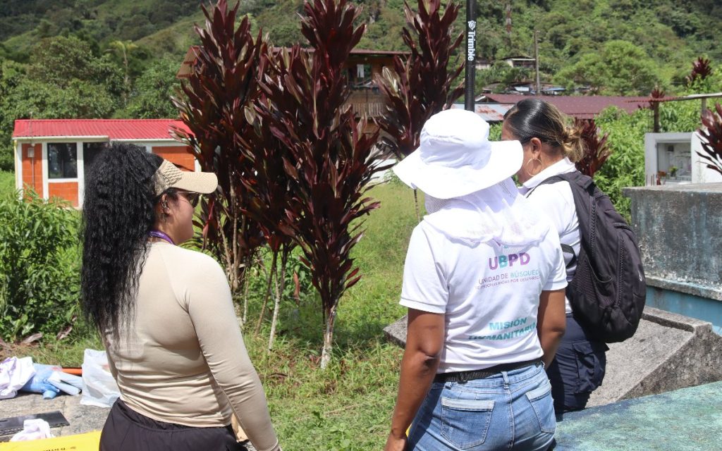 Tres investigadoras vistas de espaldas y de perfil dialogan en un entorno rural. Dos de ellas llevan camisetas blancas impresas con el logo institucional de la UBPD y viseras o sombreros para defenderse del sol. De fondo destacan plantas decorativas de hojas rojas y pequeñas construcciones.