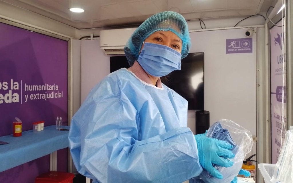 Mujer con indumentaria médica de bioseguridad, que incluye bata azul, gorro, mascarilla y guantes, sosteniendo una bolsa plástica transparente dentro de un espacio de atención. De fondo hay un pendón morado con el texto "humanitaria y extrajudicial".