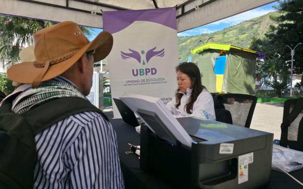 Funcionaria sentada en un puesto de atención al aire libre usando un computador portátil, hablando con un hombre con sombrero que está de espaldas. Al lado de la funcionaria hay una impresora y detrás de ella un pendón con el logotipo de la UBPD, Unidad de Búsqueda de Personas dadas por Desaparecidas.