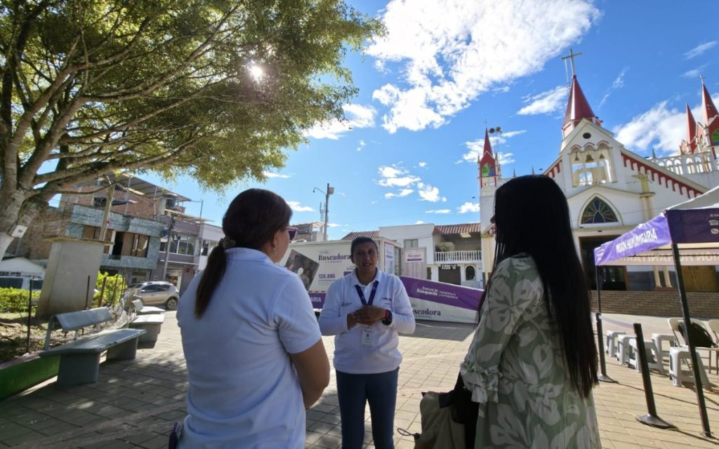 Tres mujeres conversando en la plaza principal de un municipio en un día soleado. Una de ellas, con camisa blanca e identificación corporativa, habla frente a las otras dos que están de espaldas. Al fondo se ve una carpa de atención con el letrero "Misión Humanitaria" y una iglesia blanca con detalles rojos.