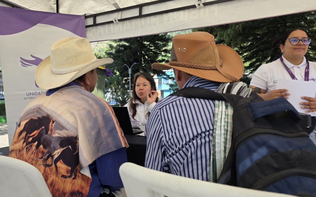 Dos funcionarias de la UBPD atendiendo a dos hombres con sombrero y ponchos tradicionales que están sentados de espaldas. Una funcionaria está sentada hablando con ellos frente a un computador, mientras que la otra está de pie a la derecha sosteniendo unos documentos.