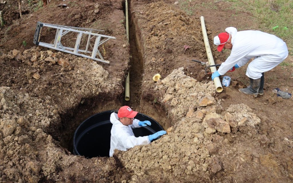Fotografía de una zona rural en tierra firme donde se realiza una excavación. Un trabajador con traje de bioseguridad blanco y gorra roja del Cruz Roja está agachado dentro de un hueco profundo instalando una estructura circular negra. A la derecha, otro trabajador con el mismo atuendo y guantes azules corta un tubo de plástico con una sierra de mano. Una zanja estrecha atraviesa el suelo y al fondo se observa una escalera metálica apoyada sobre la tierra.