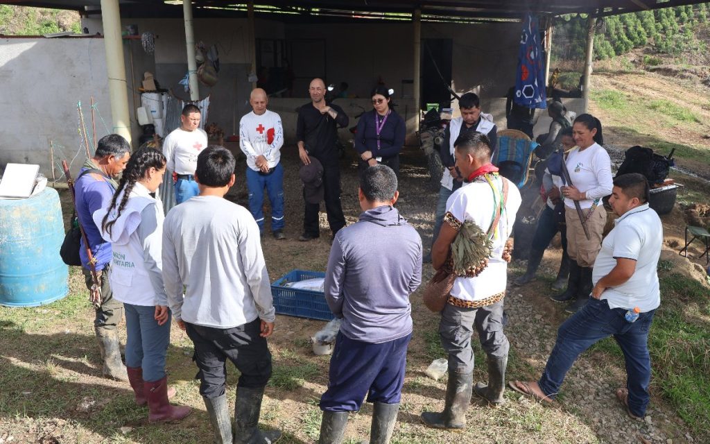 Fotografía que muestra a un grupo de personas dispuestas en círculo en una zona exterior semiabierta bajo un techo de lámina. Entre los asistentes hay miembros del pueblo indígena Awá portando indumentaria tradicional y collares, acompañados por funcionarios humanitarios con chalecos blancos y ropa táctica. En el centro del círculo, sobre el piso de tierra, hay canastillas y recipientes preparados para una ceremonia o reunión de coordinación.