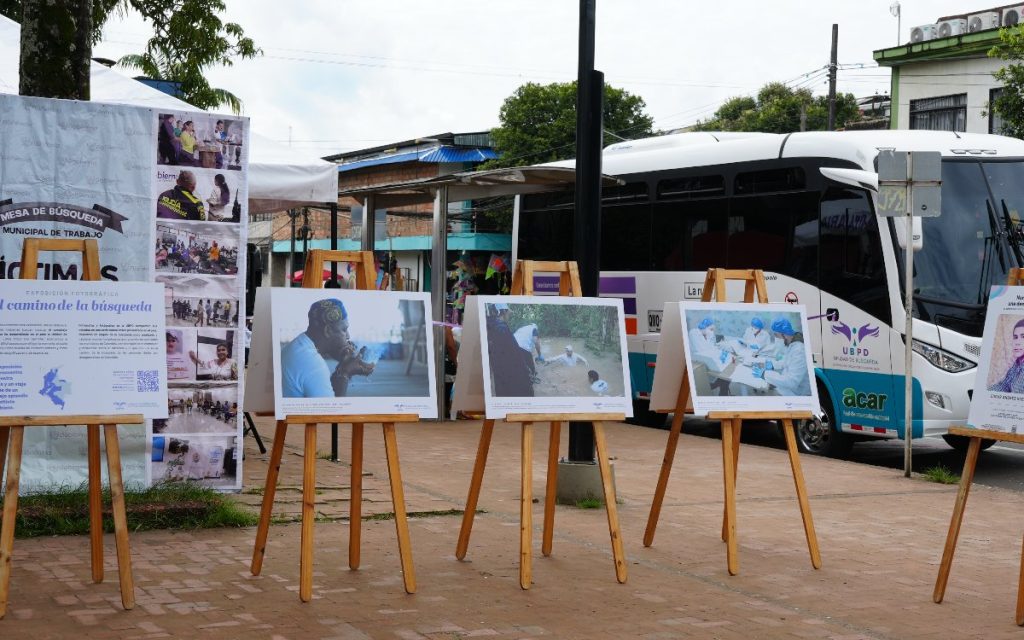 Exposición fotográfica al aire libre llamada "El camino de la búsqueda" exhibida sobre caballetes de madera en el espacio público. Al fondo se encuentra estacionado el bus de la Unidad de Búsqueda de Personas dadas por Desaparecidas (UBPD).