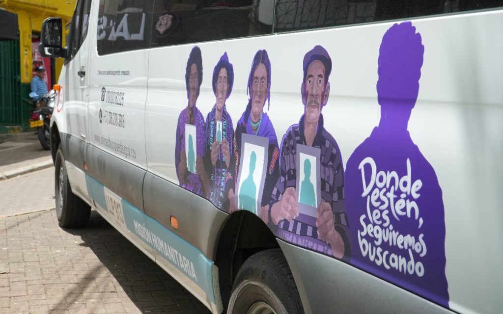 Detalle lateral del vehículo de la UBPD con una ilustración gráfica de cuatro personas que sostienen retratos de sus familiares. A la derecha, una silueta morada contiene el lema "Donde estén, los seguiremos buscando". En la parte inferior dice "Misión Humanitaria".