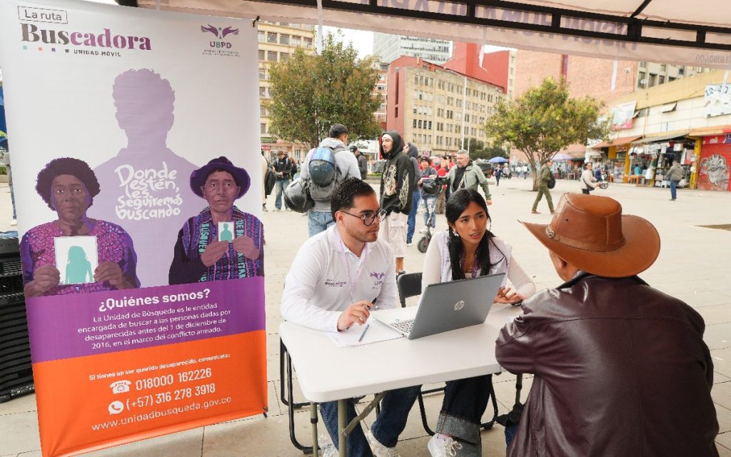 Dos funcionarios de la UBPD sentados en un escritorio con un computador portátil atienden a un ciudadano con sombrero en una carpa instalada en una plaza pública. Junto a ellos hay un pendón de "La ruta Buscadora" que incluye los números de contacto y la página web de la entidad.
