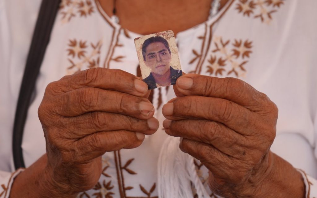 Primer plano de las manos de una persona mayor que sostiene con delicadeza la pequeña fotografía antigua de un hombre joven. La persona viste una blusa blanca con bordados marrones.