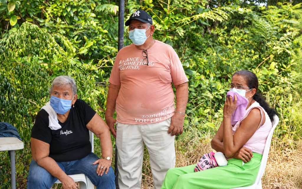 Tres personas adultas mayores permanecen sentadas y de pie al aire libre frente a vegetación verde. A la izquierda, una mujer con camiseta negra y tapabocas azul; al centro, un hombre de pie con camiseta rosada, gorra negra y tapabocas azul; a la derecha, una mujer sentada con camiseta blanca, tapabocas y una pequeña toalla morada cerca del rostro con gesto reflexivo.