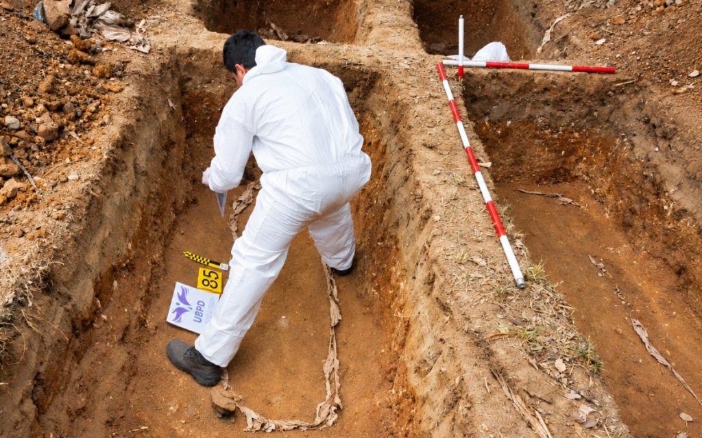 Un especialista forense con traje de bioseguridad blanco trabaja agachado dentro de una fosa rectangular. A su lado se observan reglillas de escala de medición y un letrero de señalización forense con las siglas UBPD y el número 85, además de balizas de medición rojas y blancas en el borde del terreno excavado.