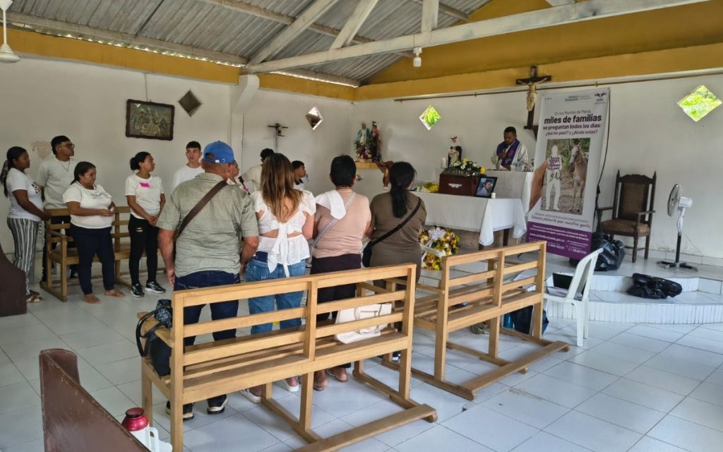 Fotografía tomada desde la parte trasera de una capilla modesta de paredes blancas. Un grupo de personas permanece de pie observando hacia el altar, donde un sacerdote oficia una misa frente a un pequeño cofre fúnebre rodeado de flores y una fotografía. Al lado del altar se observa un pendón de la Unidad de Búsqueda de Personas dadas por Desaparecidas (UBPD).