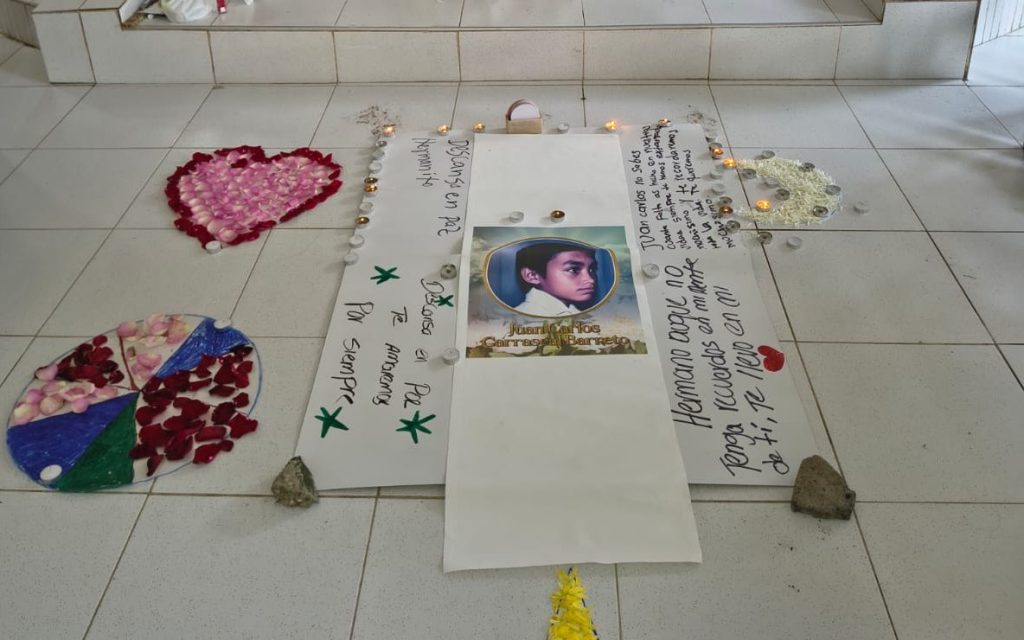Plano cenital sobre el piso de baldosas blancas de una capilla. En el centro se encuentra la fotografía de un niño con el nombre "Juan Carlos Carrascal Barreto". A su alrededor hay carteles con mensajes escritos a mano como "Descansa en paz" y "Por siempre te amaremos", acompañados por un corazón formado con pétalos rosados, velas encendidas y arreglos florales.