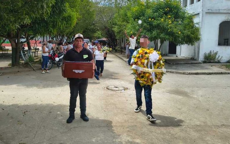 Fotografía en una calle pavimentada rodeada de árboles. Un hombre con gorra sostiene al frente el cofre fúnebre de madera, mientras a su lado otro hombre camina llevando una gran corona de flores amarillas y blancas. Al fondo se observa a un grupo de personas acompañando el recorrido.