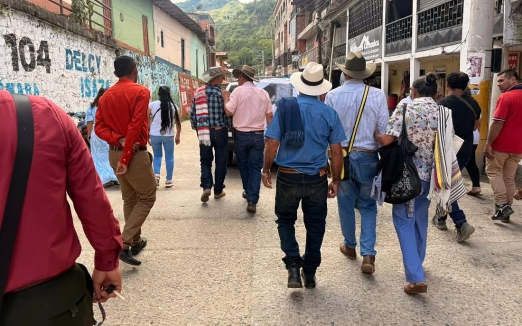 Vista trasera de un grupo de hombres y mujeres caminando por la calle de un municipio. Varios de los hombres llevan sombreros campesinos de ala ancha, camisas de botones y mochilas o mantas al hombro. Al fondo se aprecian fachadas de casas de varios pisos y colinas verdes.