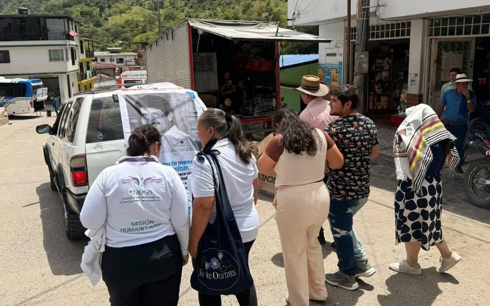 Un grupo de personas observa la parte trasera de un vehículo tipo camper blanco estacionado en una calle. En el panorámico trasero del carro hay un cartel con la fotografía de un hombre sonriente y el texto "Honramos tu memoria". Dos mujeres en primer plano llevan camisetas y bolsos con los logos institucional de la Unidad de Búsqueda de Personas dadas por Desaparecidas (UBPD) y la frase "Misión Humanitaria".