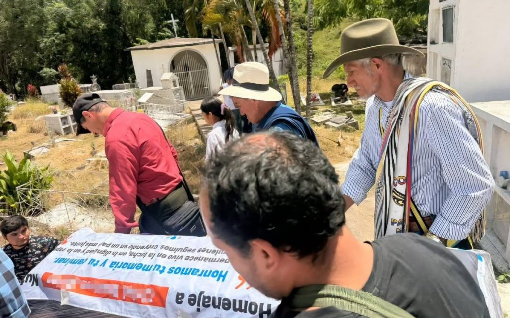 Plano picado en un cementerio al aire libre donde varias personas, entre ellas campesinos con sombreros tradicionales y mochilas tejidas, sostienen y observan una pancarta con el mensaje: "Honramos tu memoria y tu caminar. Permanece vivo en la lucha, en la dignidad y en la esperanza...". De fondo se ven bóvedas blancas y vegetación.